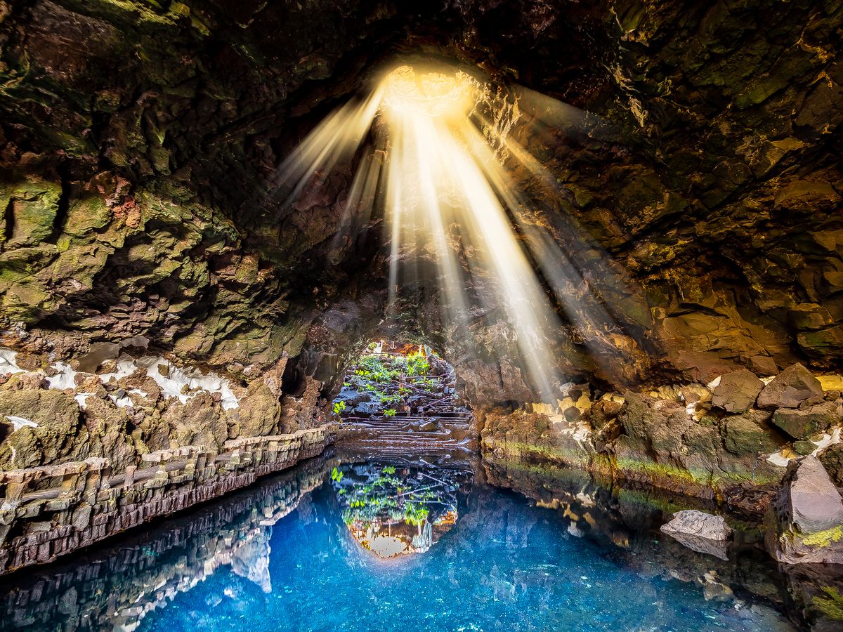 Bilden visar grottlandskapet Jameos del Agua i Timanfaya nationalpark. Här syns det stilla vattnet i mitten av bilden, omgivet av grottväggar. Solljuset lyser ner i grottan och vattnet från mitten av taket