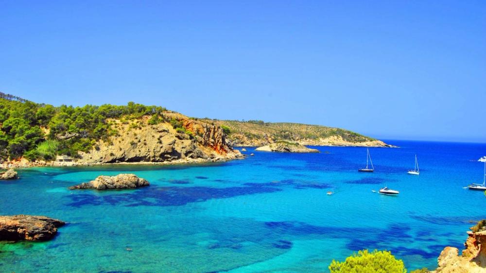 Krystallklart blått hav ved en steinete kyst på Ibiza, med seilbåter på det turkise vannet og grønne åser i bakgrunnen.