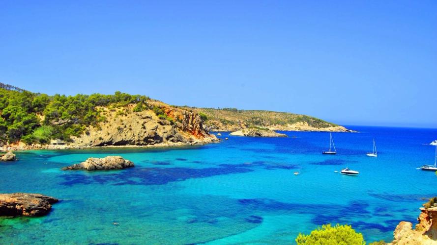 Krystallklart blått hav ved en steinete kyst på Ibiza, med seilbåter på det turkise vannet og grønne åser i bakgrunnen.