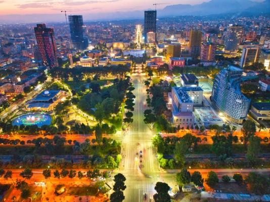 Bildet viser sentrum av Tirana i Albania tatt med drone på kveldstid. I midten en bredere og opplyst trafikkaveny, med by-begyggelse på hver side. Mellom bebyggelsen en del frodig beplantning.