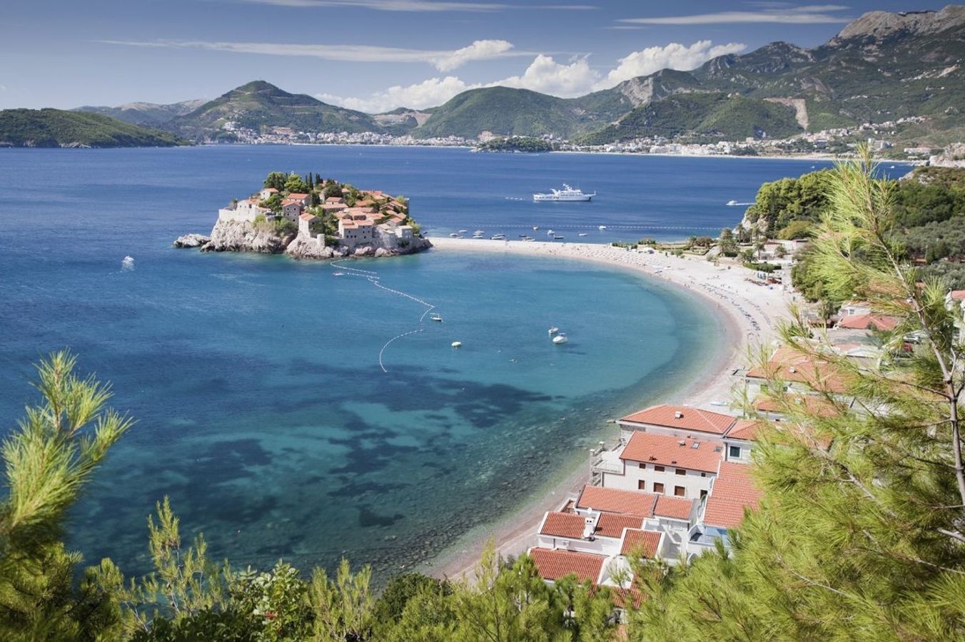 Panoramautsikt over kysten ved Sveti Stefan. I vannet ligger den tidligere middelalderbyen på en klippeøy, forbundet med fastlandet via stranden. Langs det turkisblå havet strekker stranden seg, og i bakgrunnen reiser fjellene seg.
