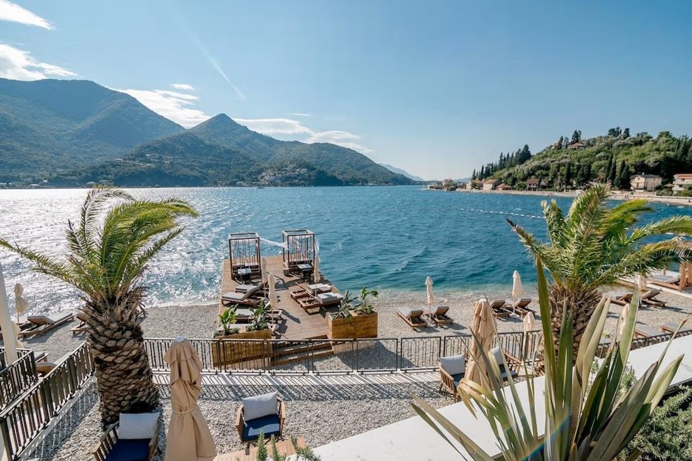 Bilden visar utsikten från Du hittar hotellet Boutique Hotel Casa Del Mare Mediterraneo mellan Tivat och Herceg Novi i Montenegro. Det blå havet med en stenig strandkant, palmer, solstolar och en liten privat brygga. Längst in i bilden ser man ett frodigt bergslandskap.