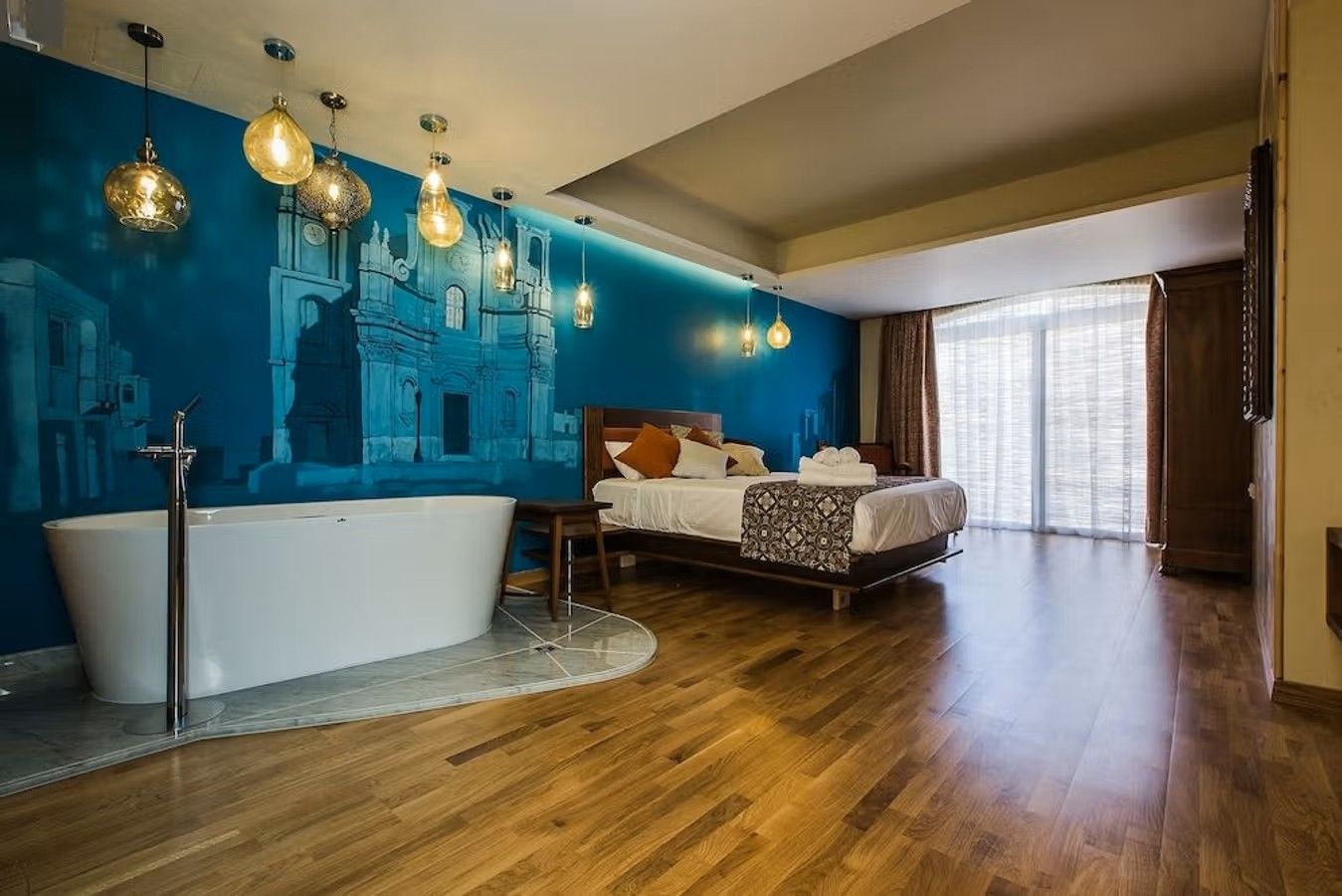 Rummen på det familjeägda hotellet Murella Living på ön Gozo på Malta har alla en unik design. Här ser vi ett större rum med parkettgolv, en dubbelsäng mitt i rummet framför en stor blå vägg. I den öppna planlösningen finns det även ett badkar.