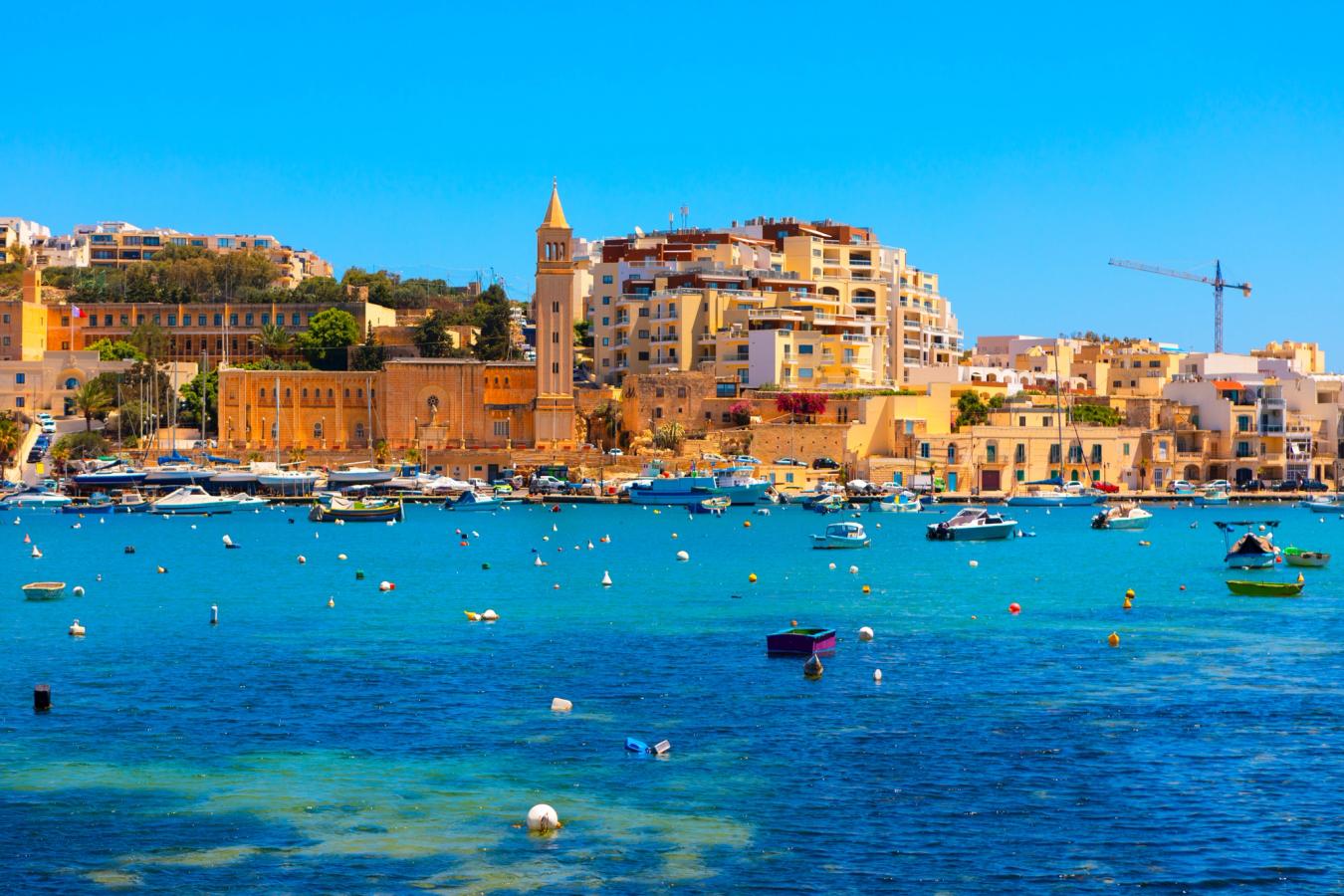 Blåt hav med både i havnen i Marsakala med byen i baggrunden. 