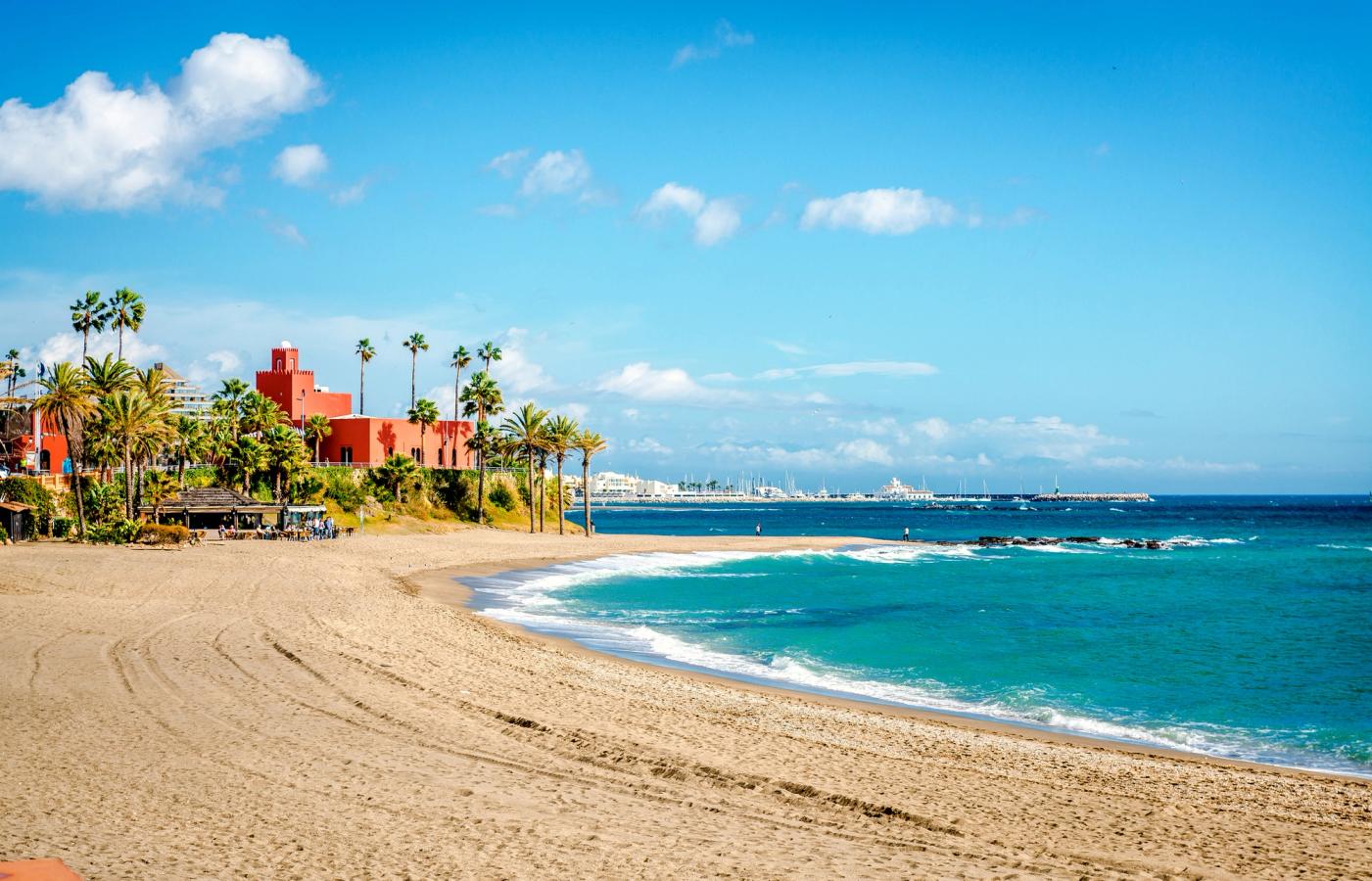Bildet viser en nypreparert strand i Benalmadena. Fremst i bildet selve stranda og vannet som sklir innover sanda. Lengr einn i bildet, på venstre side ser vi en taverna med besøk og deler av et hotell rett ovenfor. Lengst inn i bildet ser man konturene av en marina. Alt under en nesten skyfri himmel.