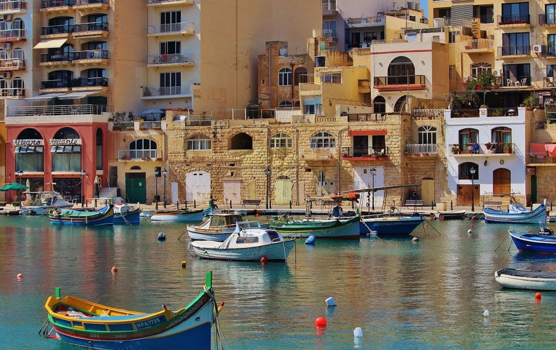 Färgglada båtar i en hamn på Malta, med traditionella byggnader och moderna lägenhetskomplex i bakgrunden.
