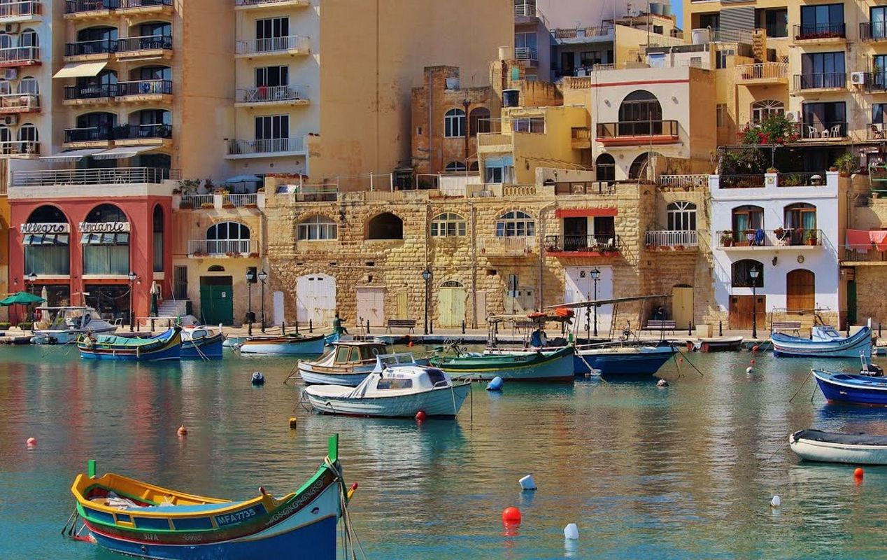 Farverige både i en havn på Malta, med traditionelle bygninger og moderne lejlighedskomplekser i baggrunden.
