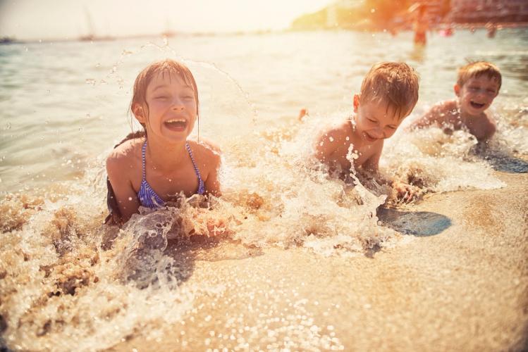 3 barn liggende i vannkanten på stranda, mens småbølger skyller rundt dem