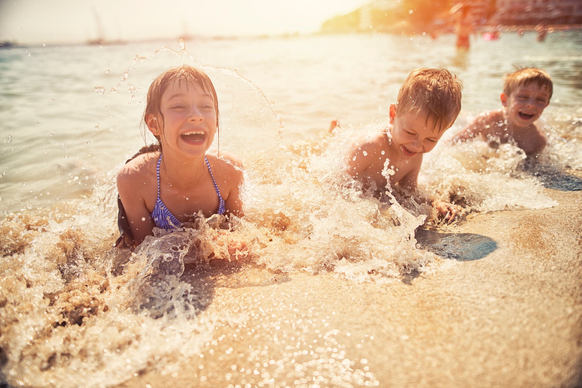 3 børn ligger i vandkanten på stranden, mens små bølger skyller ind over dem.