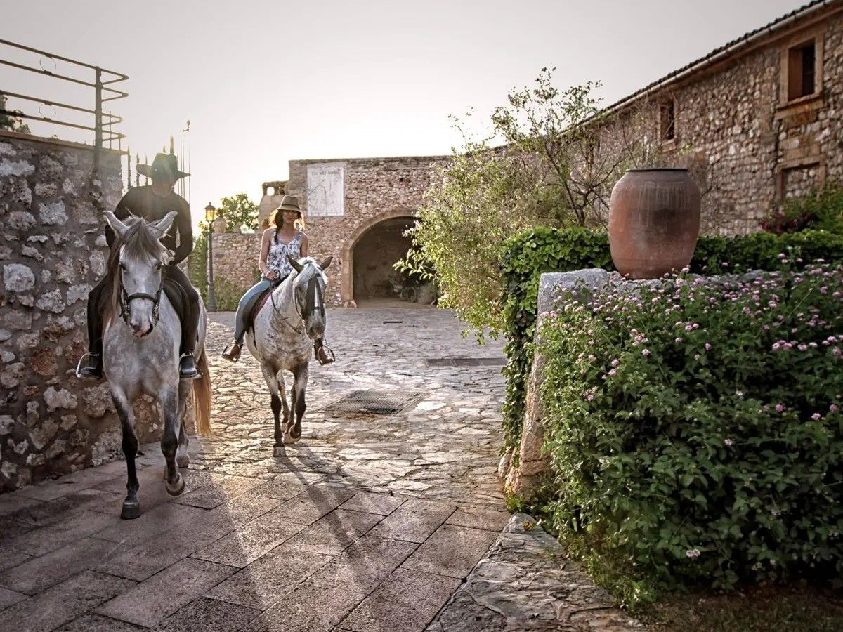 Fra gårdspladsen på hotellet Sa Bassa Rotja Ecoturisme. Husmure og mure er i sten, og i midten ser vi to personer på hver sin hest ride ud af gårdspladsen.