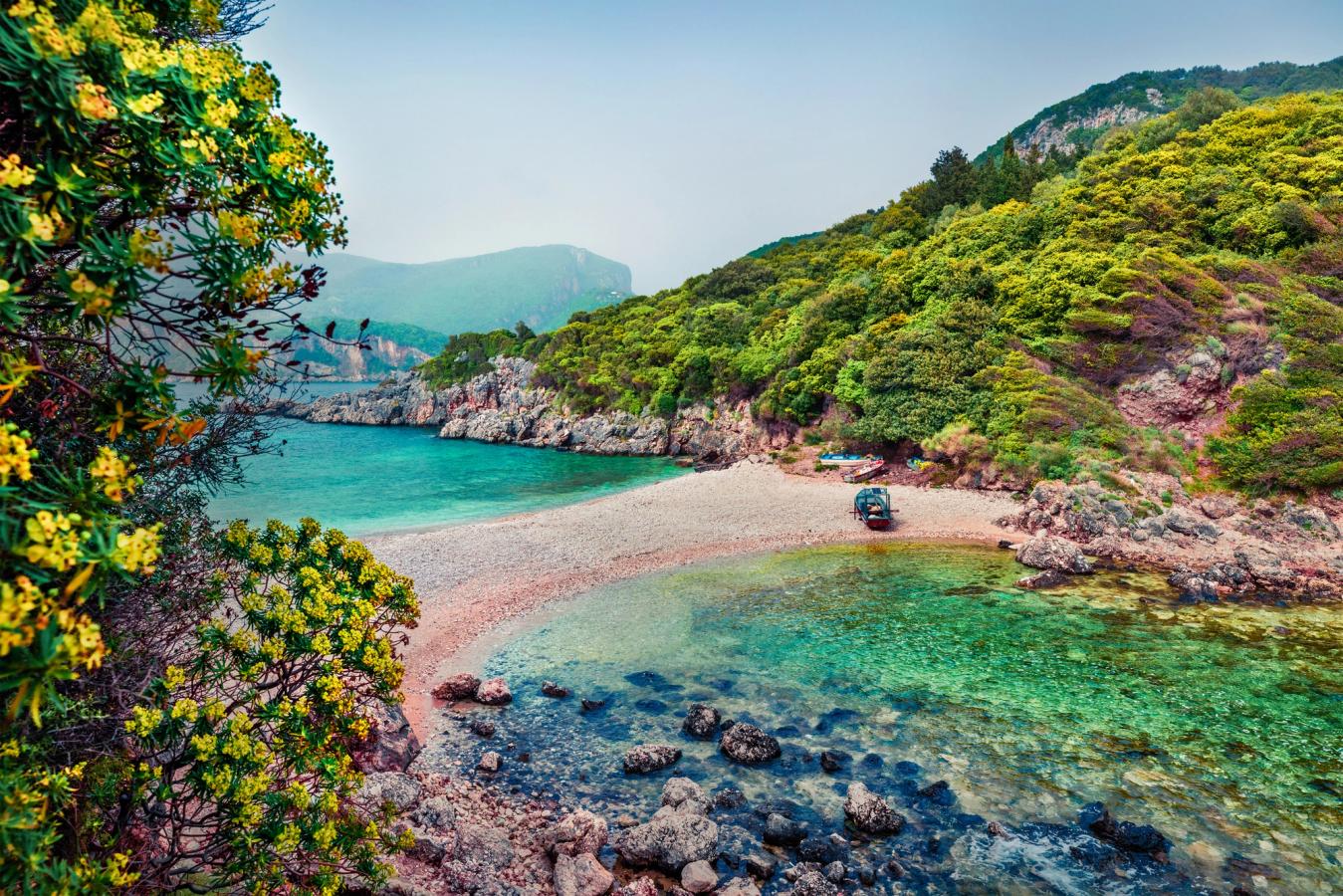 Billedet viser Limni Beach på Korfu i Grækenland. Den lille strand er omgivet af krystalklart og stillestående vand på begge sider. I hver ende af stranden er der et frodigt naturlandskab og en skyfri himmel.