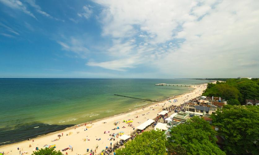 Billedet viser den lange strand langs Kołobrzeg og Østersøen i Polen. Stranden med havet til venstre og naturen med nogle bygninger til højre i billedet.