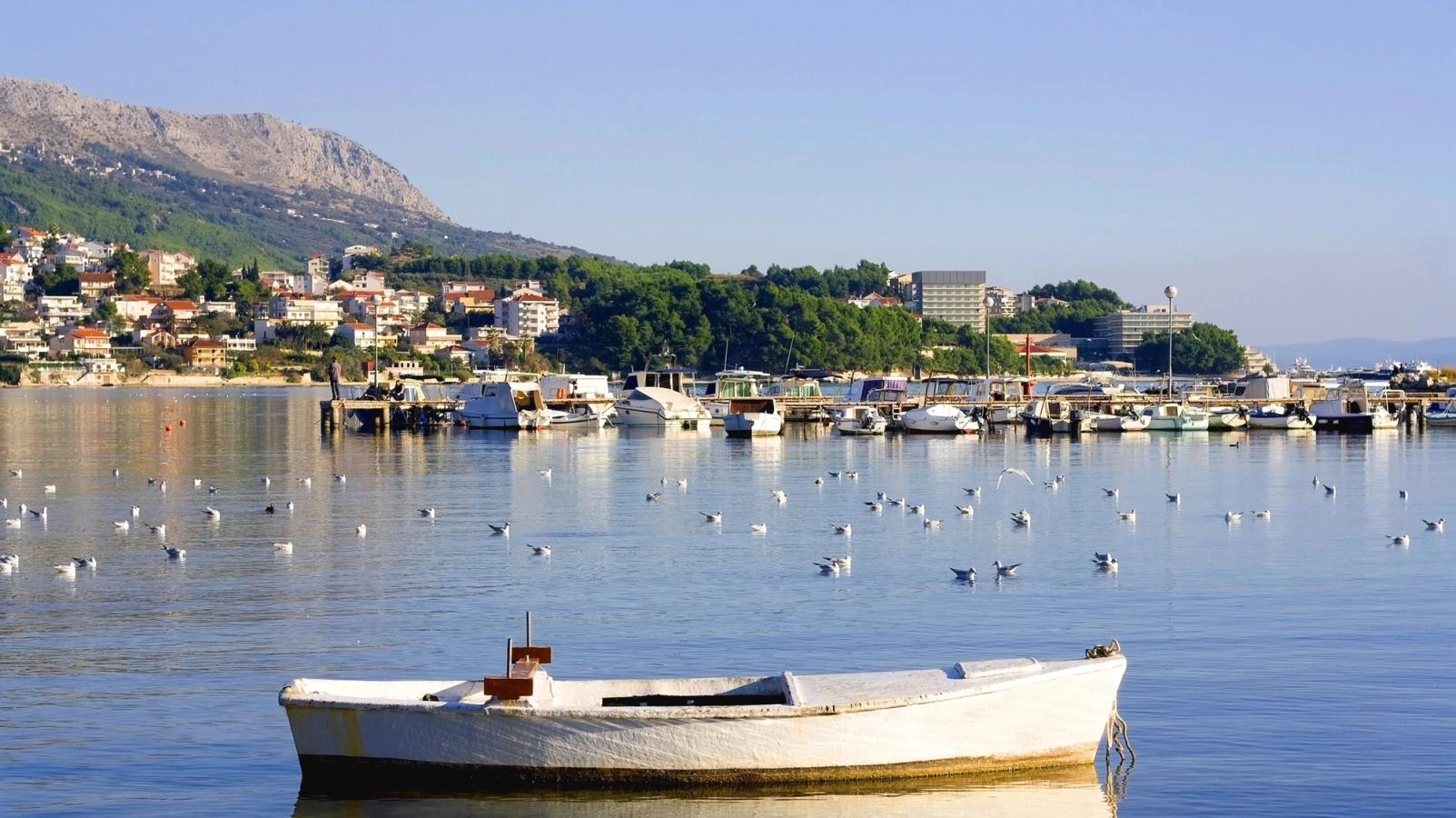 En liten vit båt flyter stilla på vattnet i en bukt omgiven av måsar. I bakgrunden syns en marina med många båtar, en kuststad med röda tegeltak, gröna träd och berg i horisonten under en klarblå himmel.