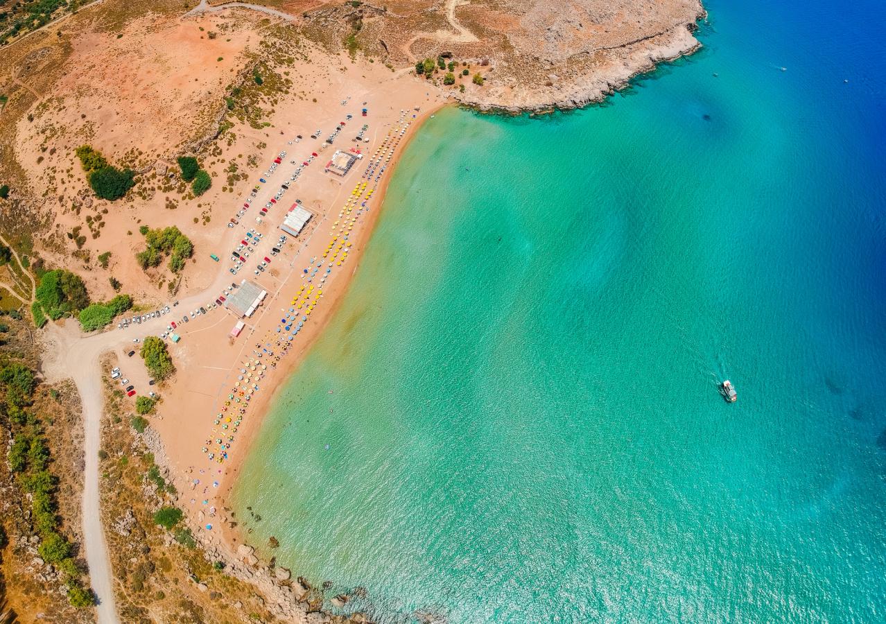 Bilden visar ett drönarfoto över Agia Agathi Beach. Till höger syns det lugna och kristallklara vattnet, i en färgkombination av blått och grönt. Till vänster i bilden syns den breda stranden med badgäster och bakom dem bilar på en parkeringsplats