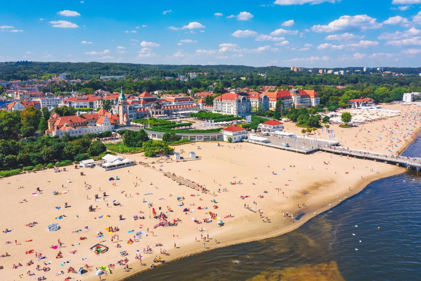 Billedet viser den lange og brede strand i Sopot, fyldt med badegæster. Bag stranden ses Sheraton Sopot Hotel centralt i billedet, omgivet af bygninger med røde tage og frodig beplantning.