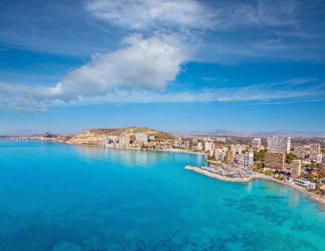 Strandbilden visar Playa Albufereta i Alicante med gyllene sandstrand, blått hav och vita byggnader i bakgrunden under en klar, solig himmel.