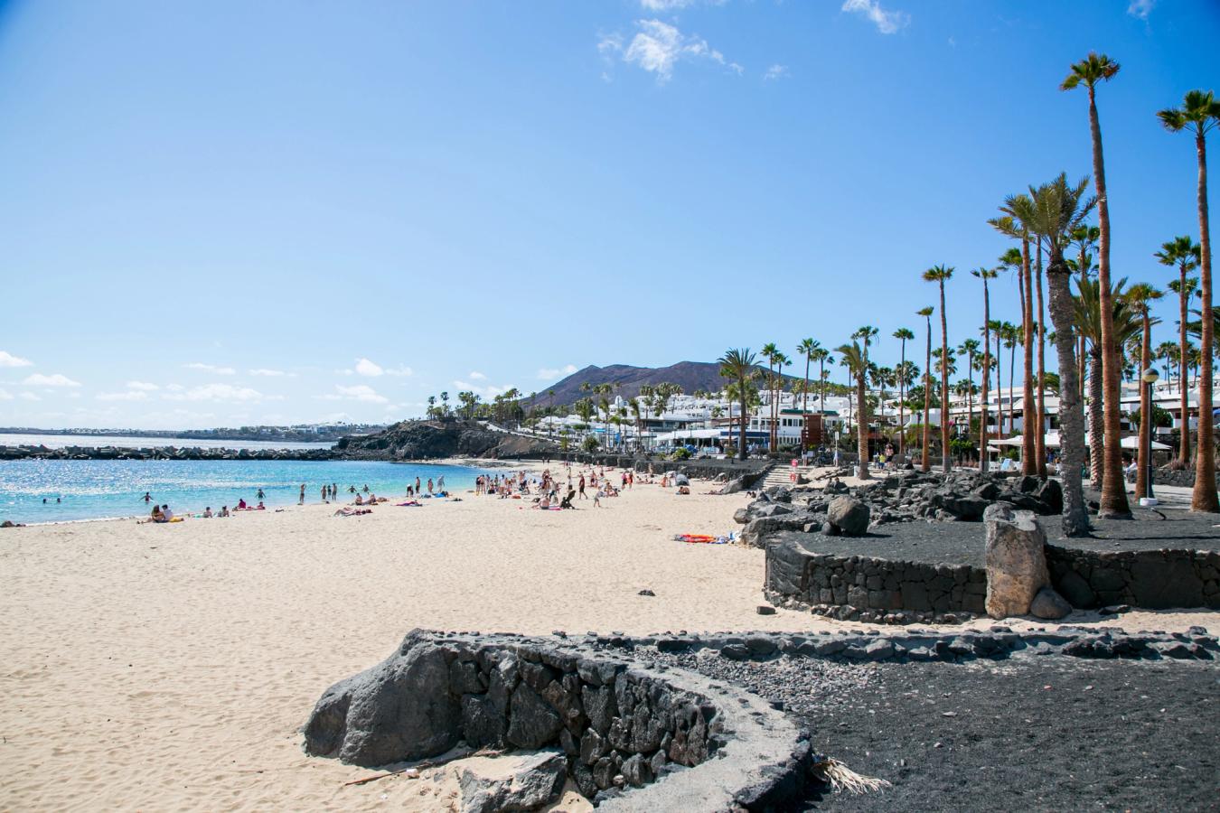Bilden visar stranden Playa Flamingo, precis intill staden Playa Blanca. I förgrunden ser man några svarta lavformationer som smälter samman med en lång strand och havet på vänster sida av bilden. I fjärran skymtar man staden Playa Blanca