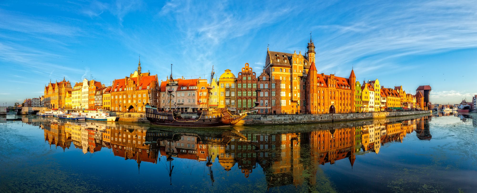 Bild av bebyggelsen i Gdansk längs flodstranden. I floden speglar sig stadsbilden med en nästan molnfri himmel.