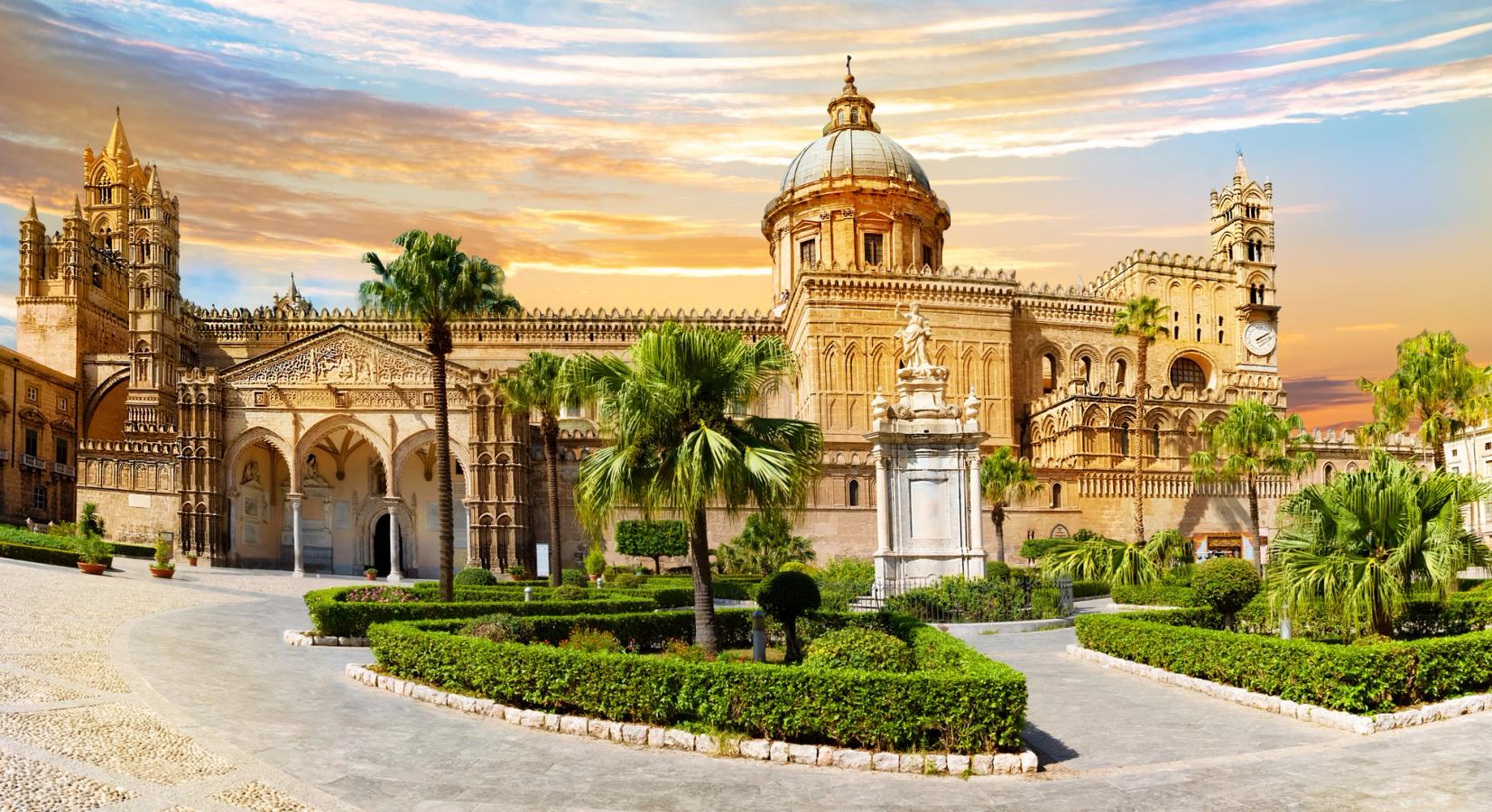 Bildet er av fremsiden på katedralen i Palermo med kupler og spir. Foran en beplantet hage under en påstartet kveldssol