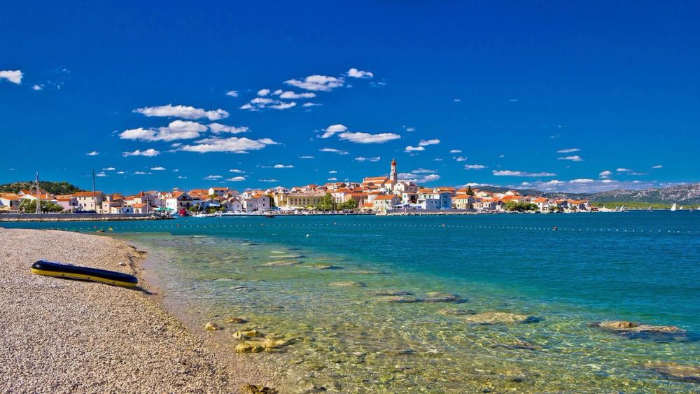 Vakker stenstrand å bade på med kristallklart vann og utsikt til den lille byen med hvite hus og kirker bak havnen.