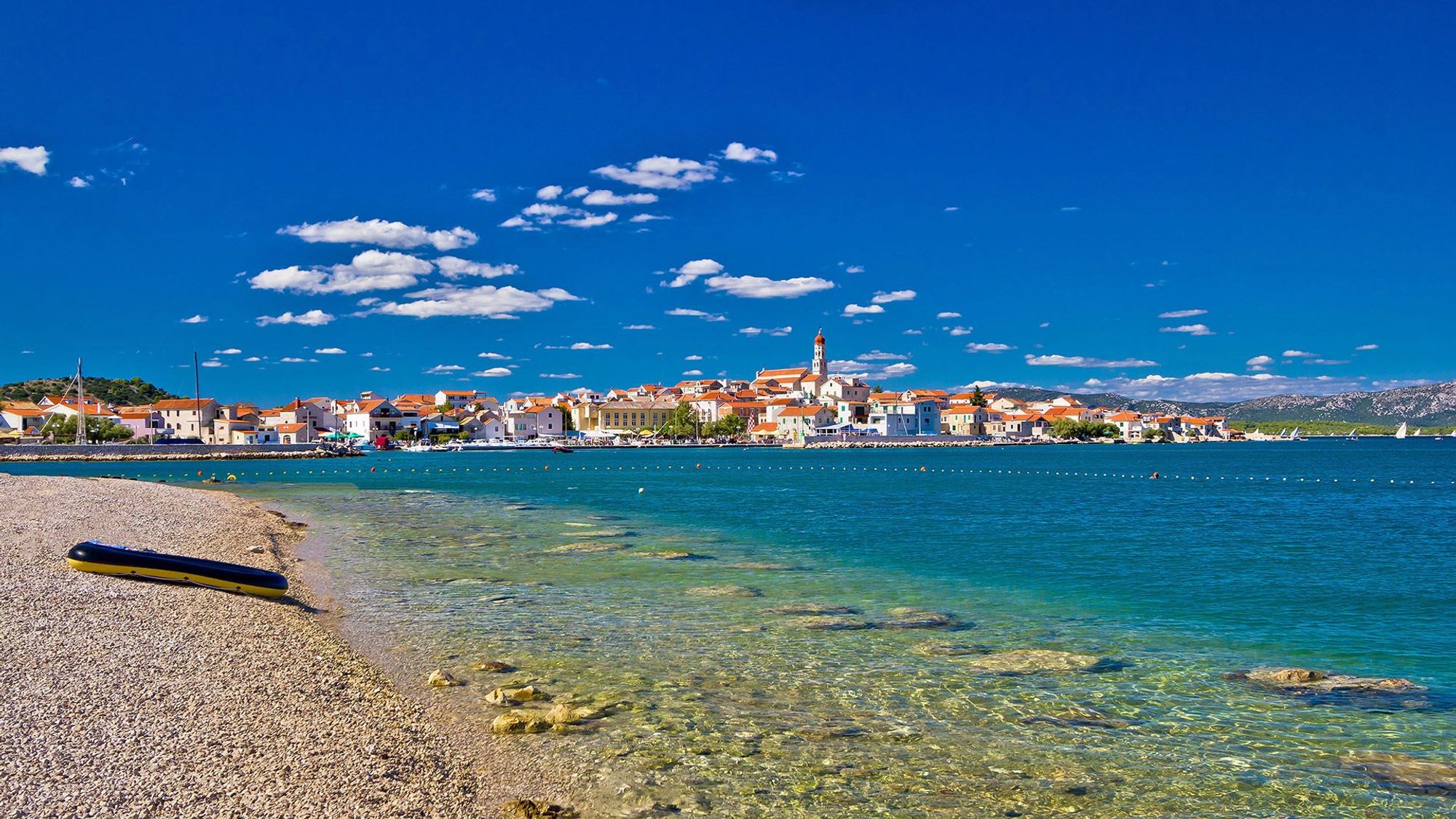 Smuk stenstrand at bade ved med krystalklart vand og udsigt til den lille by med hvide huse og kirker bag havnen.