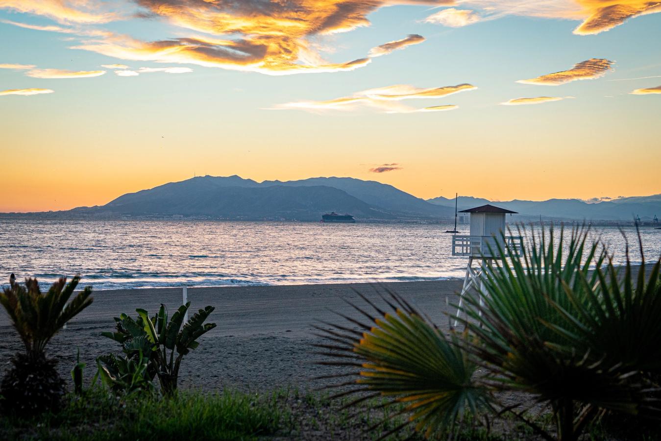 Bildet viser stranden i Rincón de la Victoria ved solnedgang. I forgrunnen ses noen tropiske planter, og i bakgrunnen ses fjellene