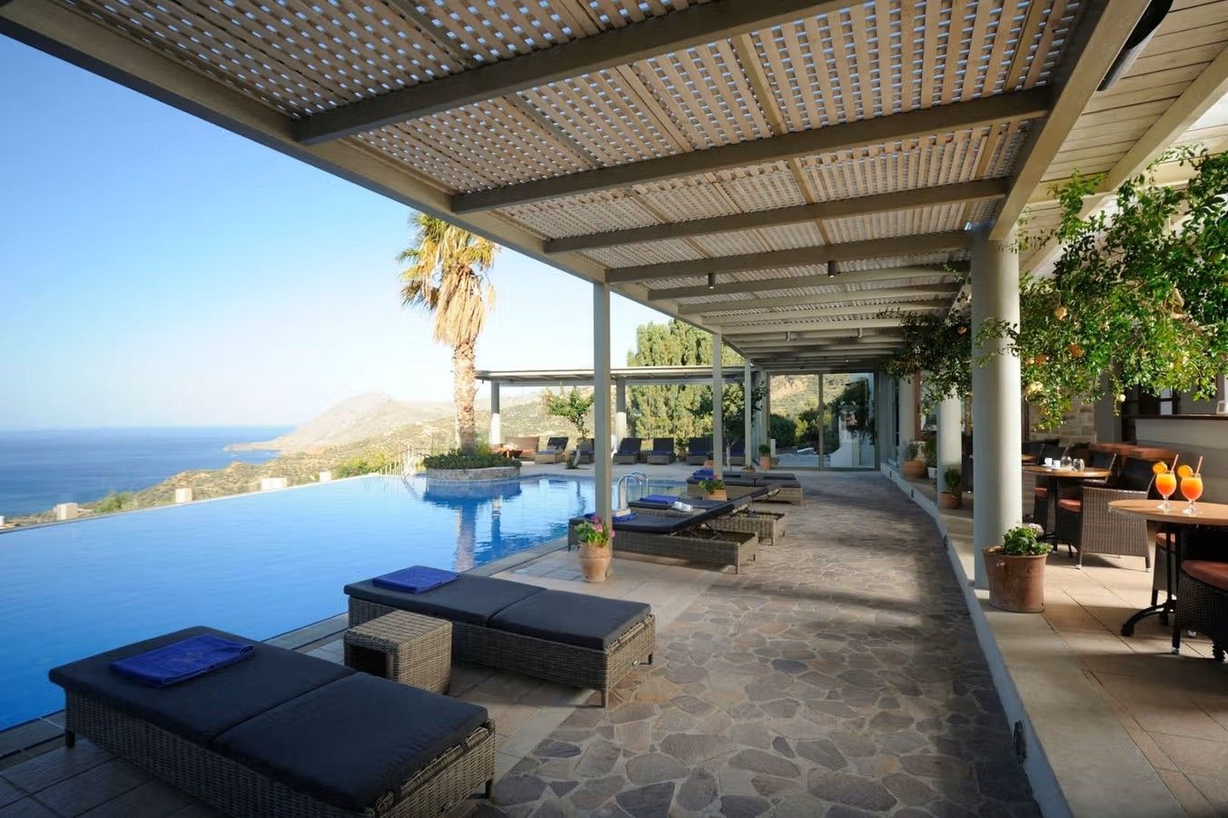 Bilden visar en infinitypool på hotellet Stefanos Village på Kreta. Från poolen har man direkt havsutsikt. Längs poolen står det solstolar där vissa är placerade under tak. Längst till höger står kafébord.