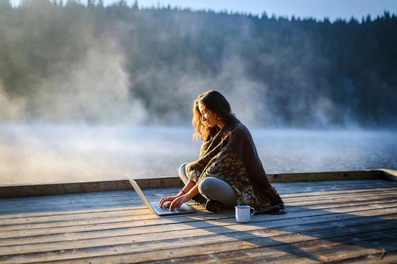 En kvinna sitter på en träbrygga med sin bärbara dator och en kaffekopp. Över vattnet ligger en låg och genomskinlig dimma