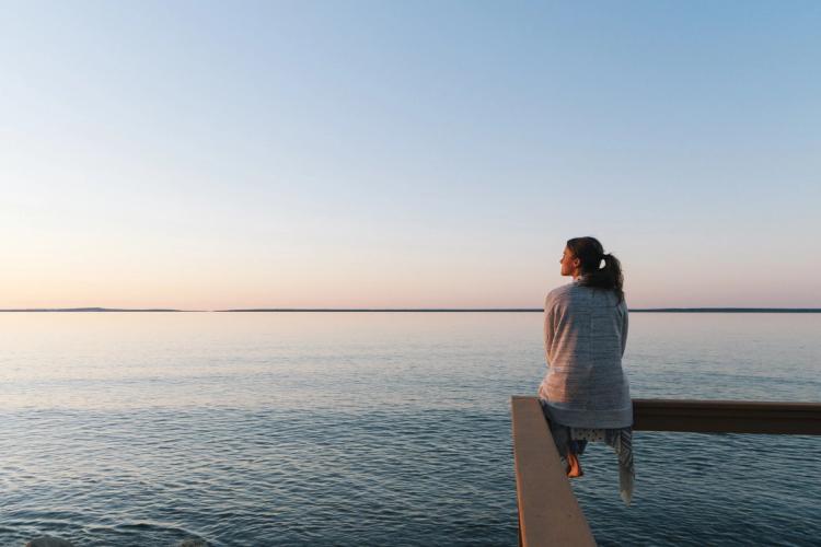 En kvinna med hästsvans sitter på kanten av en träbrygga och dinglar med benen, medan hon blickar ut över havet
