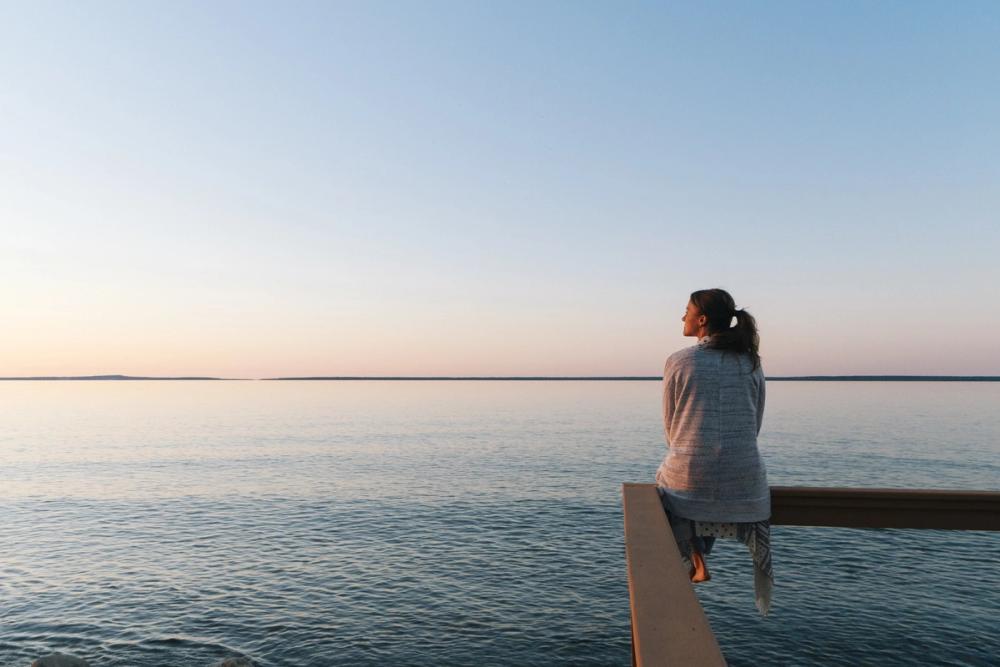 En kvinna med hästsvans sitter på kanten av en träbrygga och dinglar med benen, medan hon blickar ut över havet
