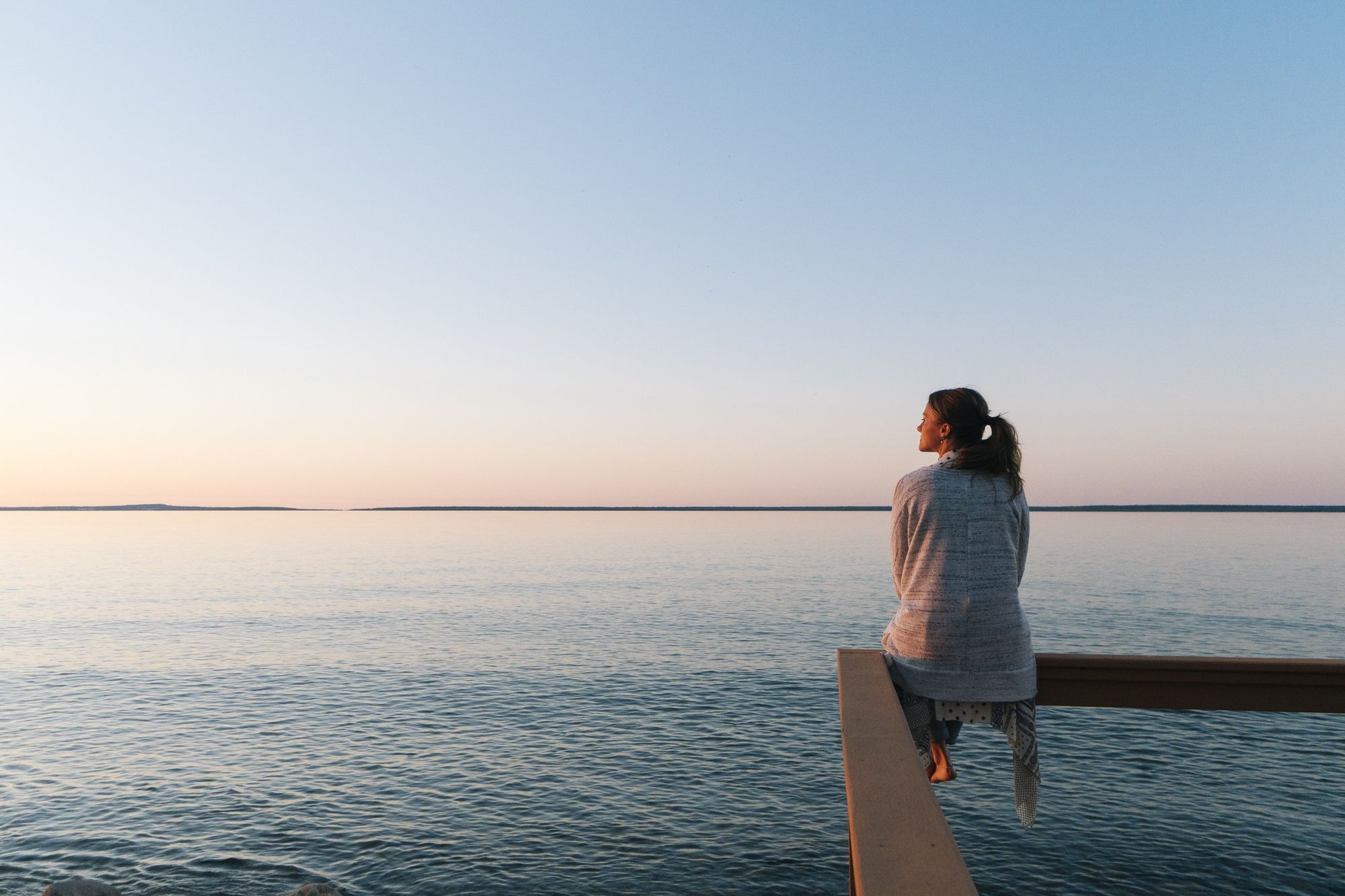 En kvinna med hästsvans sitter på kanten av en träbrygga och dinglar med benen, medan hon blickar ut över havet