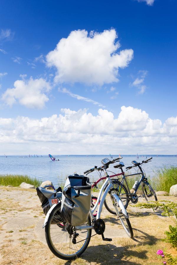 I forgrunnen står to sykler parkert mot havet, hvor flere kitesurfere driver rundt. Himmelen er blå, mens det ligger flere skyer i horisonten.