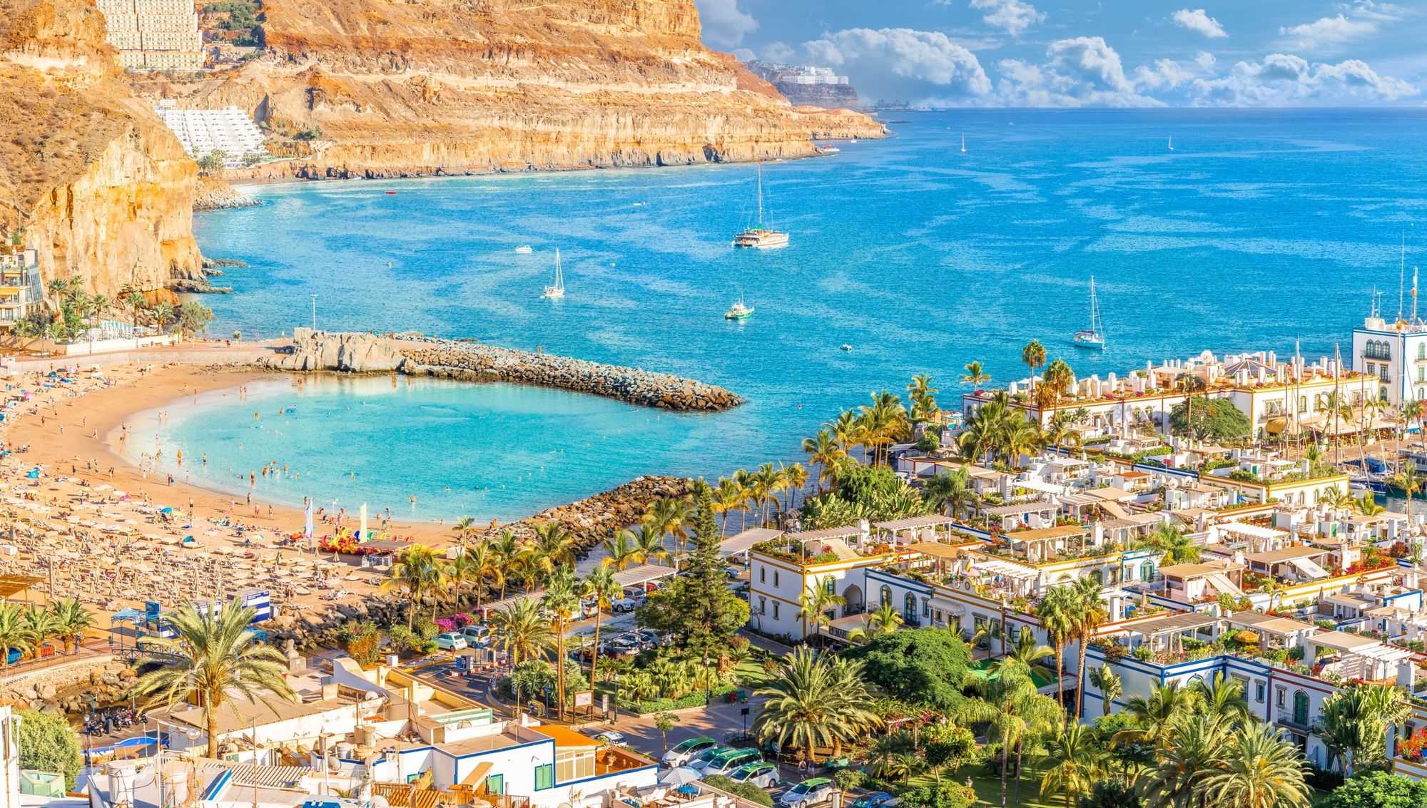 Her fra Puerto de Mogan på Gran Canaria. Vi ser den lille bystranden beskyttet bak en molo med det krystallklare og stille vannet som brer seg utover. Bebyggelsen bak stranden er lavtbygget og gir en annen dimensjon og sjarm sammenliknet med andre steder på Gran Canaria.  Rundt byen ser man større åskammer og fjellformasjoner