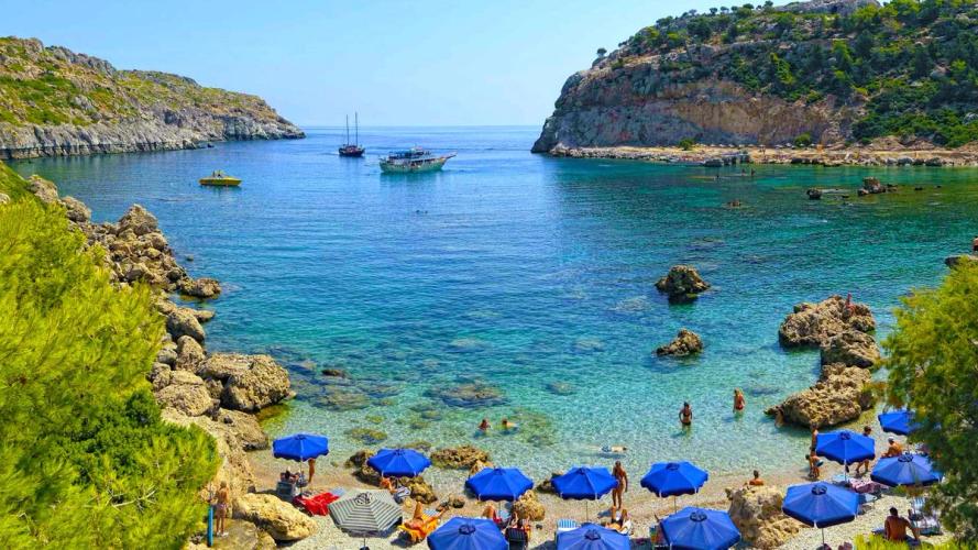 Bildet er fra populære Anthony Quinn Bay. Nærmest i bildet den smale bukta og stranden med blå parasoller og badegjester. Innover ser vi det stille og krystallklare vannet med enkelte seilbåter. På hver sin side av bukta et frodig klippeterreng.