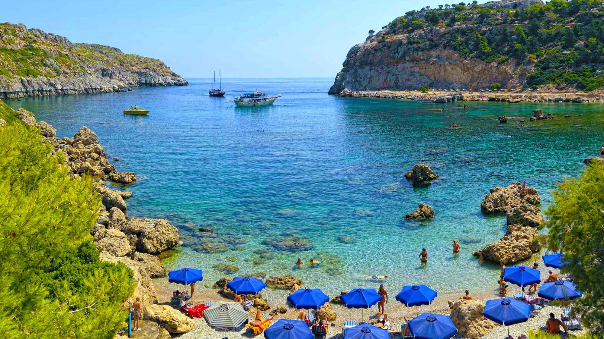 Bildet er fra populære Anthony Quinn Bay. Nærmest i bildet den smale bukta og stranden med blå parasoller og badegjester. Innover ser vi det stille og krystallklare vannet med enkelte seilbåter. På hver sin side av bukta et frodig klippeterreng.