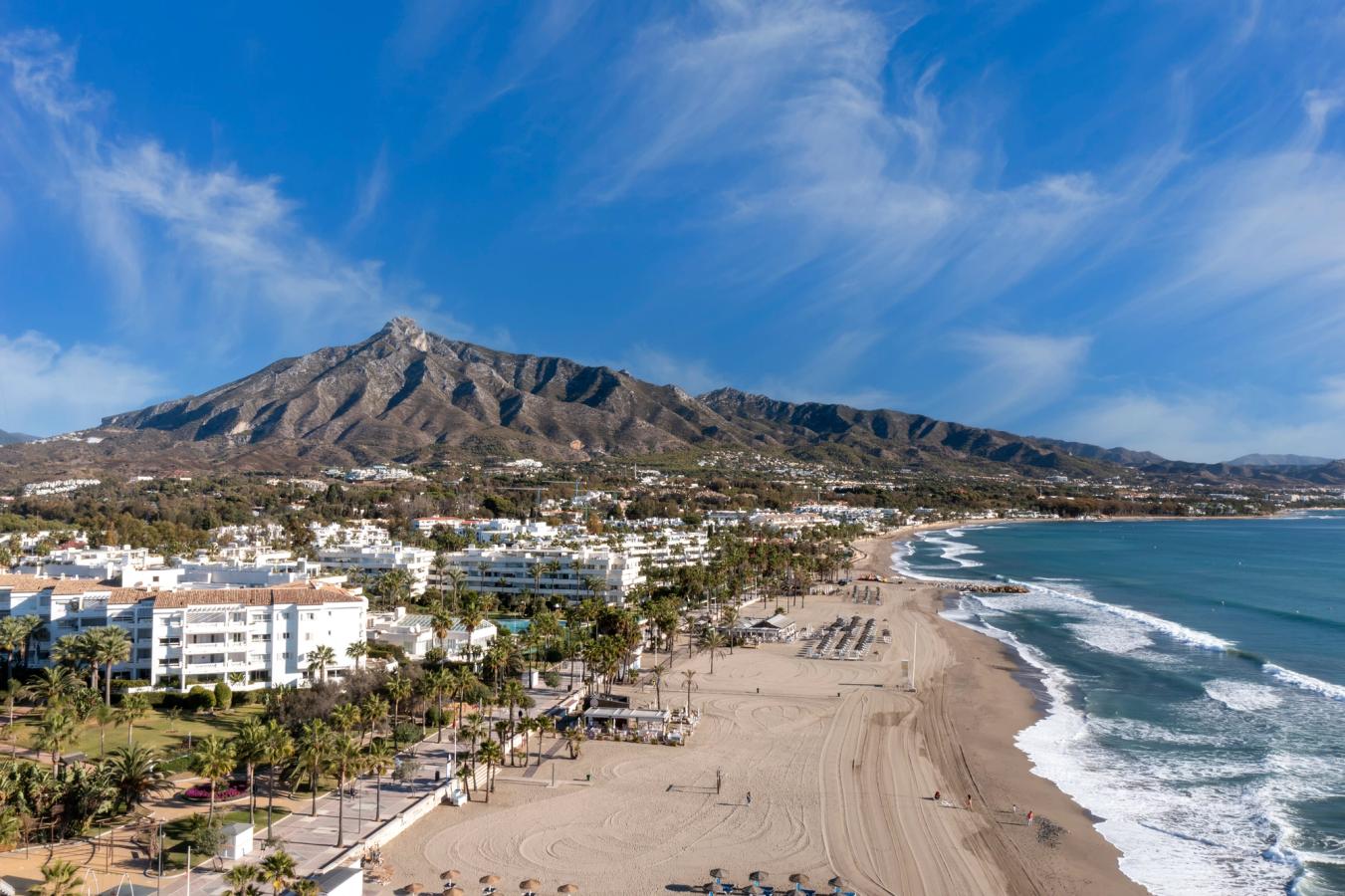 Luftfoto af sandstranden i Puerto Banus. Stranden bred og bølgerne slår ind mod kysten. Langs promenaden står grøn vegetation og her ligger også flere hoteller. 