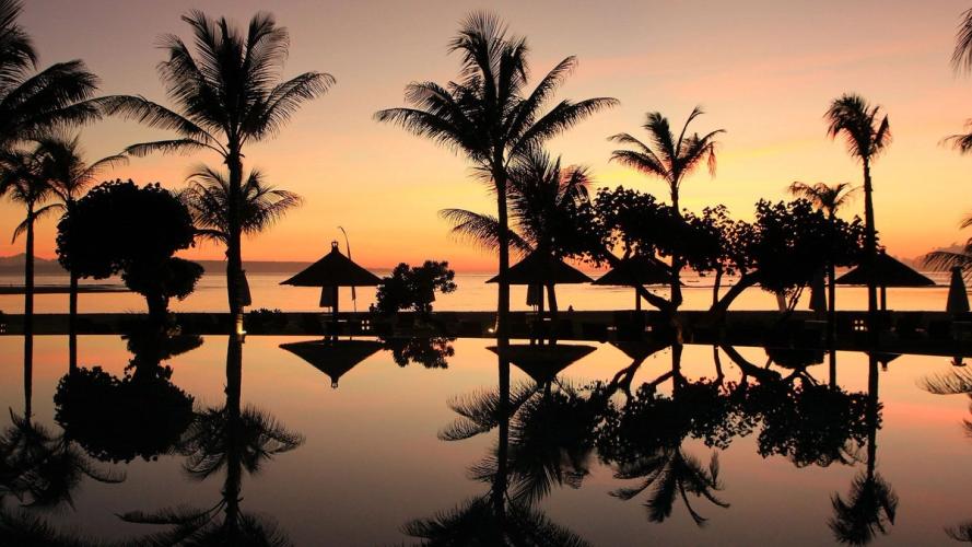 En solnedgang set fra et poolområde. Foran poolen står flere palmetræer og parasoller. Himlen er oplyst i orange, lyserøde og lilla nuancer.