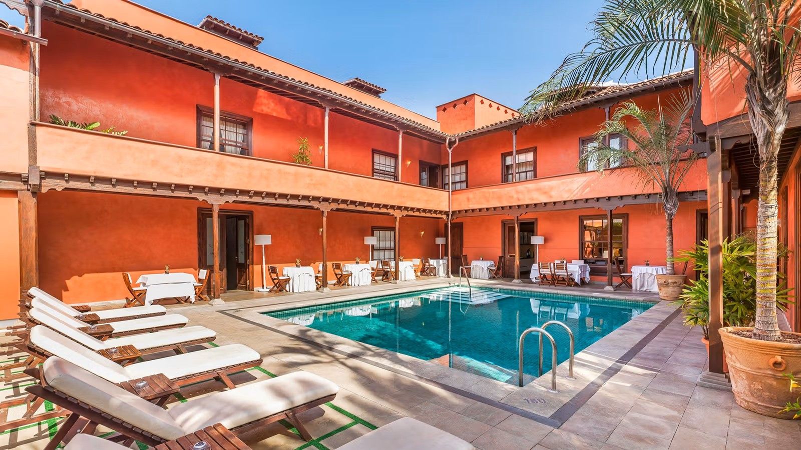 I mitten av bilden en mindre pool som först omges av solstolar och frukostbord. Därefter omges den av ett tvåvåningshotell i rödrostig färg med en genomgående balkong från ände till ände.