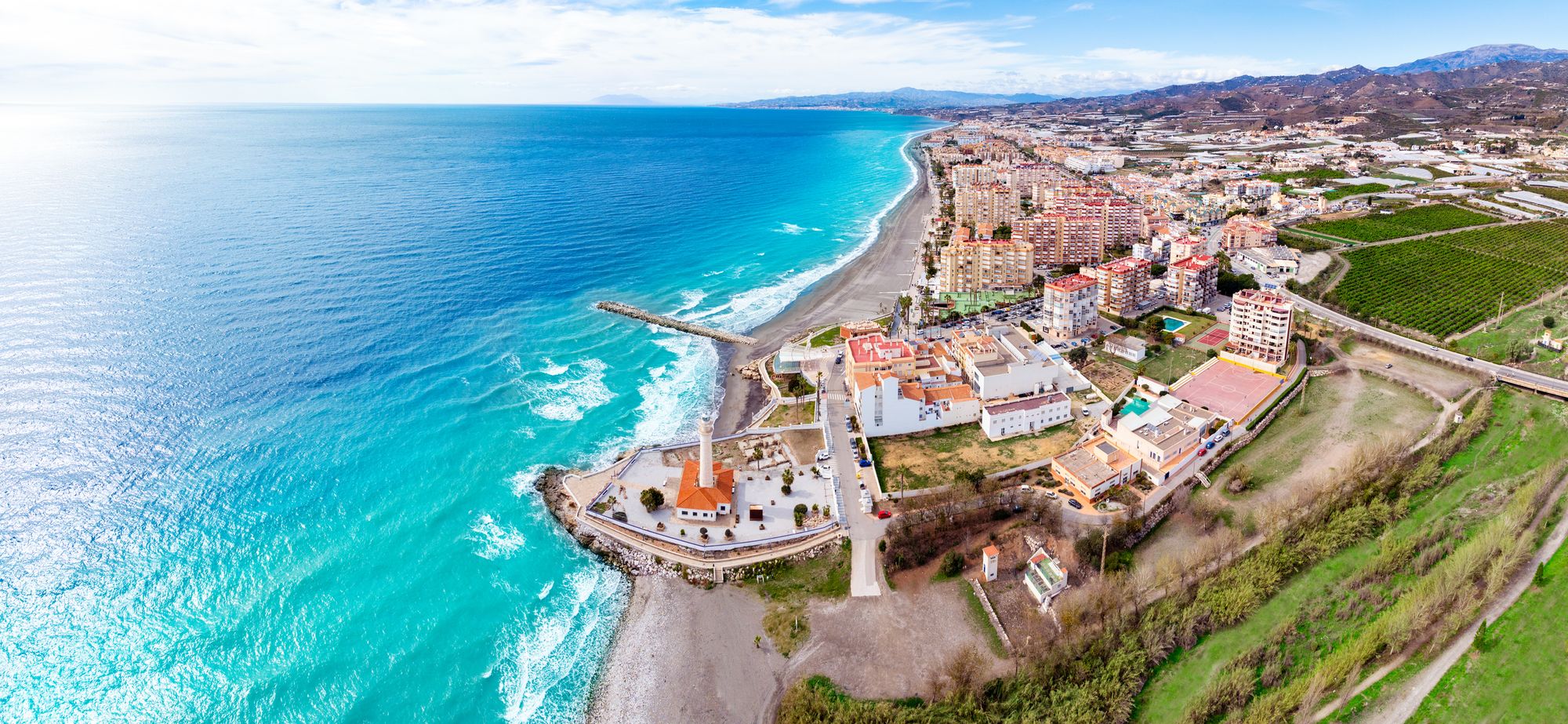 Luftfoto af Torrox Costa på Costa del Sol med det karakteristiske fyrtårn ved kysten, brede strande og det turkisblå Middelhav langs byen.
