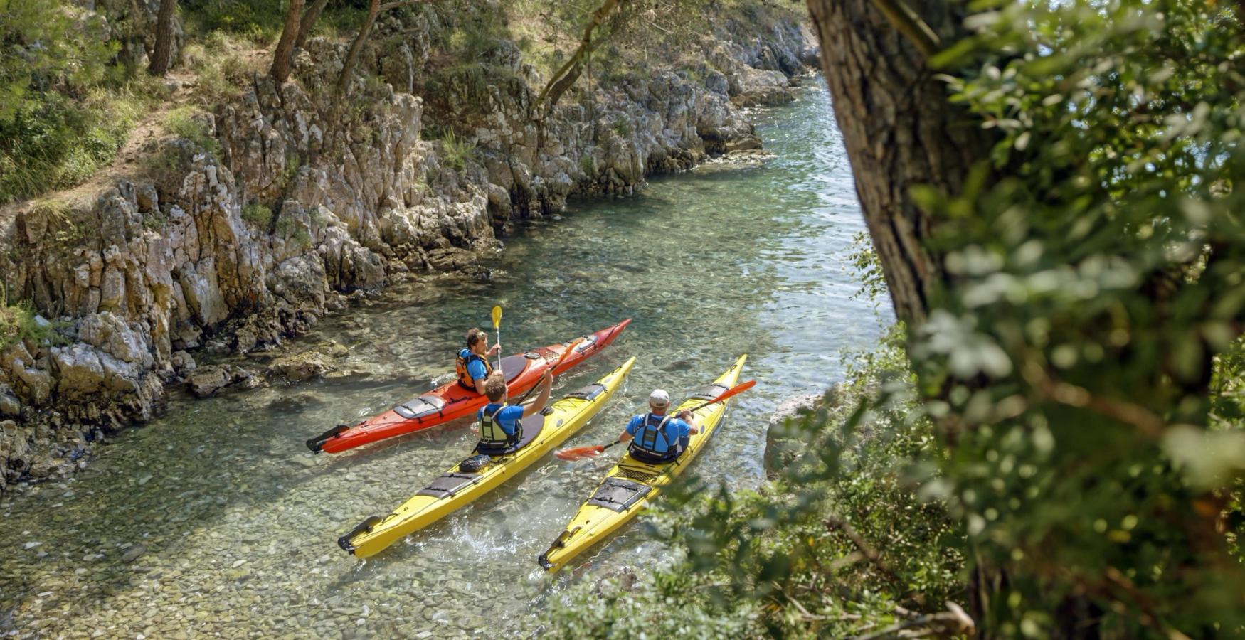 Tre personer i farverige havkajakker padler gennem en smal, lavvandet bugt omgivet af klippevægge og fyrretræer. Vandet er klart og lavt med synlige sten på bunden, og naturen omkring er frodig og grøn