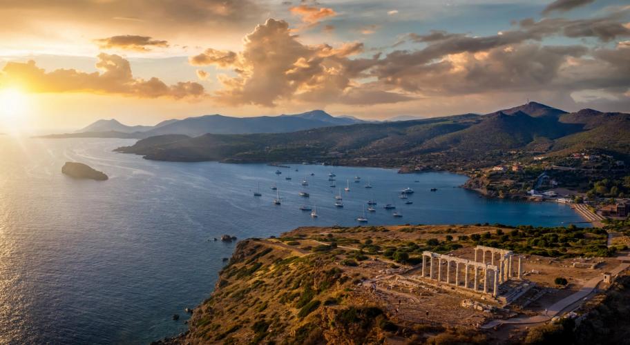 Først i bildet ser vi ruinene av Temple of Poseidon som ligger på klippeplatå med utsikt over havet. I sjøen ligger det et titallsbåter på blikkstille hav. Himmelen er småskyet i gulaktig solnedgang