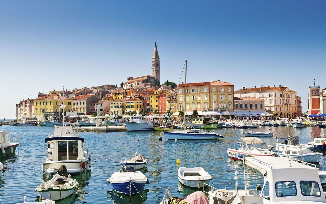 Fargerik havnepromenade i Rovinj, Kroatia. I forgrunnen ligger små båter i en rolig havn. Bak dem sees pastellfargede hus, kafeer og et høyt kirkespir som stikker opp over byens tak. Himmelen er blå og skyfri.