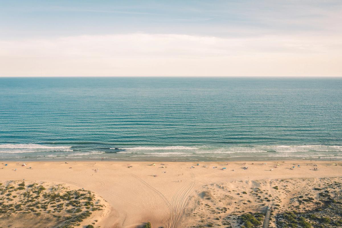 Turkosgröna vågor rullar in mot den breda sandstranden Cabanas Beach. Längs stranden finns människor som njuter av solen, och små dyner med grönskande växtlighet pryder landskapet.