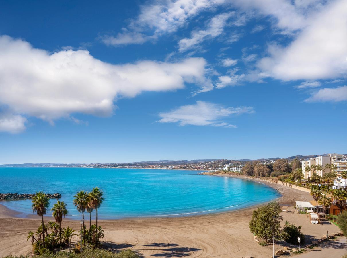 Bildet viser stranden Playa del Cristo beach ved byen Estepona. Stranden er buet som en bukt og det flotte blikkstille vannet ligger fristende midt i bildet. Stranden er noen spredte palmer og i høyre ytterkant av bildet skimter man bybildet og bebyggelse. Himmelen er blå med noen få spredte skyer.