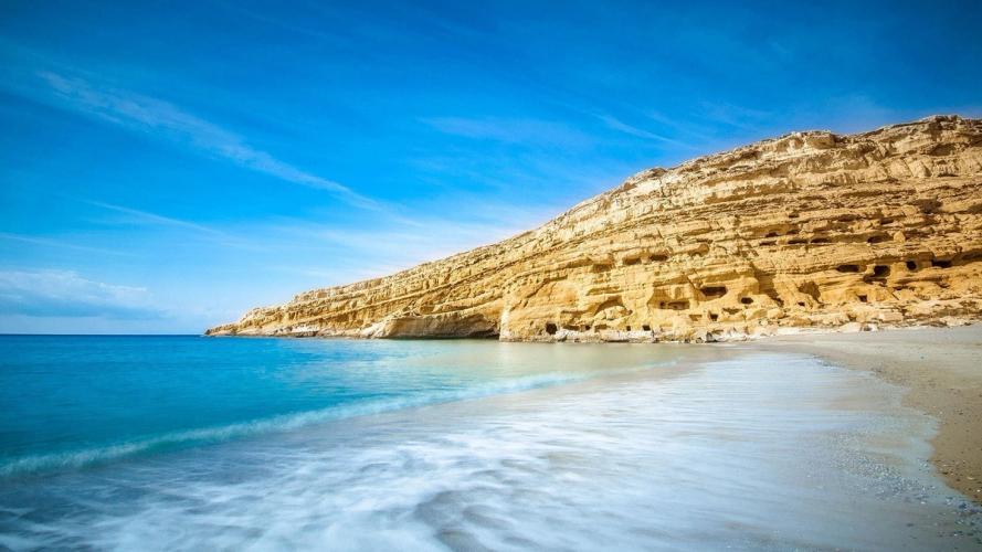 Bildet viser den vakre Matalastranden på Kreta i Hellas. Sanden viser en langgrunn og finkornet sandstrand med rolige og små bølger som duver mot strandkanten. I enden av stranden stiger det opp en klippevegg, litt kalksteinliknende.