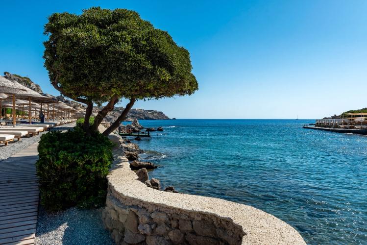 Fra Kalithea viser bildet en strandpromenade med solsenger og parasoller til venstre. Det grønnblå og krystallklare vannet i midten og til høyre en liten utstikker med ytterligere parasoller.