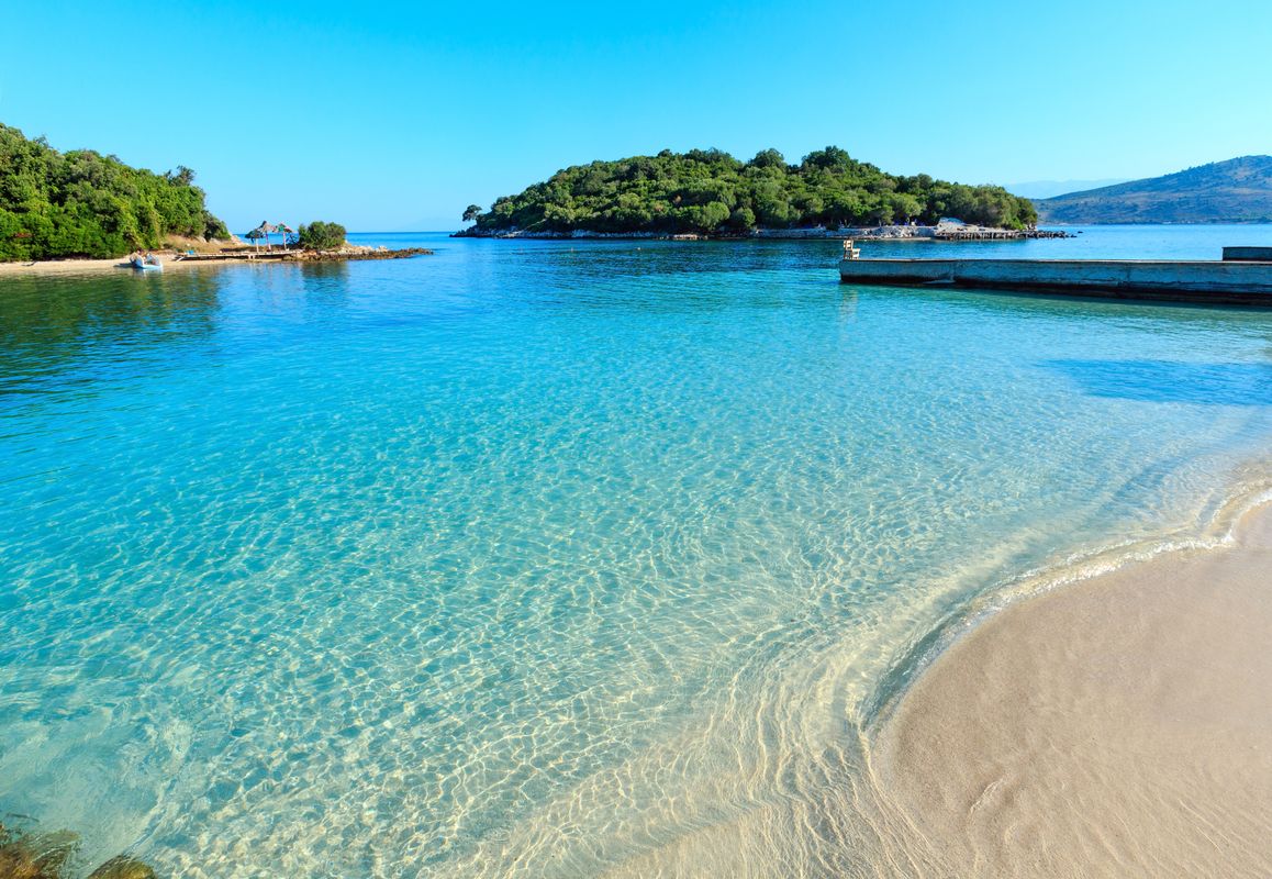 Bilden visar det stillastående blå vattnet vid stranden Ksamil Beach. Till höger ser man en bit av stranden med en smal brygga som går ut i havet. Mitt i bilden syns två små öar under en molnfri himmel