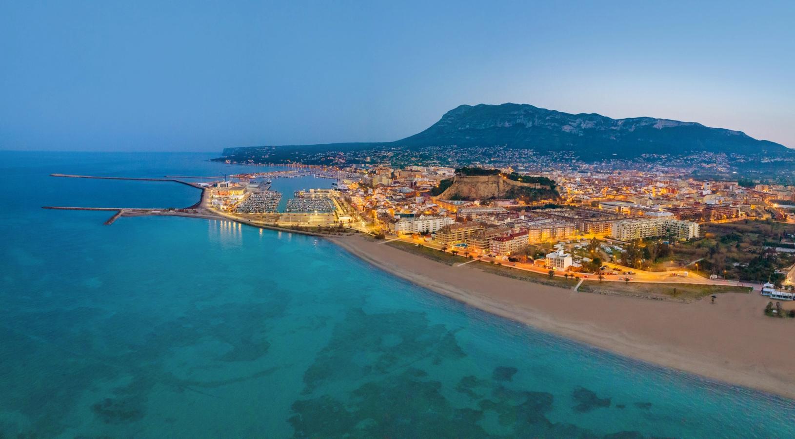 Utsikt over Denia på kveldstid. Byen lyser opp mens havet er turkisblått i skumringen. 