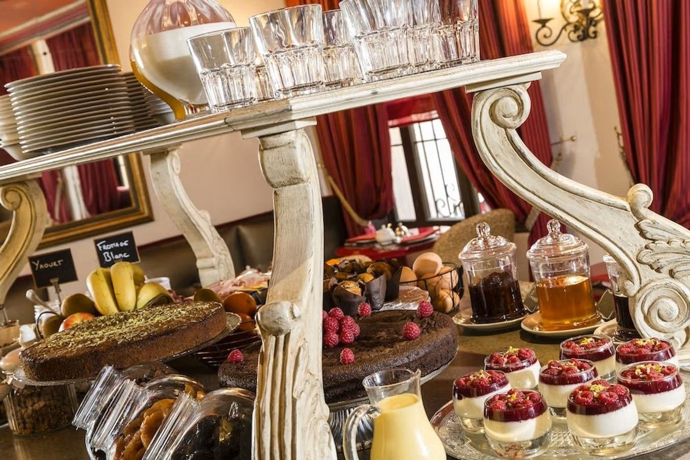 Bildet viser eget bakverk fra hotellet Le Cavendish i Cannes. Midt i bildet et hvitt serveringsbord i 2 etasjer. Nederst er fylt med mørk kake og ulike glass med youghurt, honning og sauser. Øverste delen er det stablet serveringsglass i asjetter. Lengst inn i bildet er et vindu med mørkerøde gardiner.