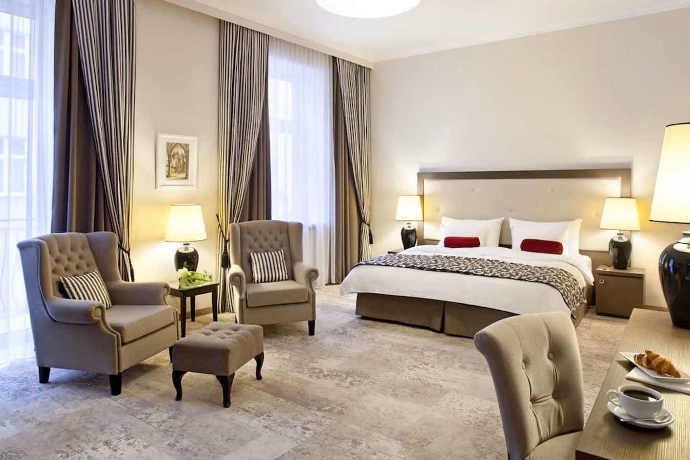 Bildet er fra en suite på Metropolitan Boutique Hotel i Krakow i Polen. Et stort luftig rom med stor takhøyde, hvor dobbeltsengen står inntil en vegg midt i rommet. Det står plassert to komfortable lenestoler til venstre ved to høye vinduer.